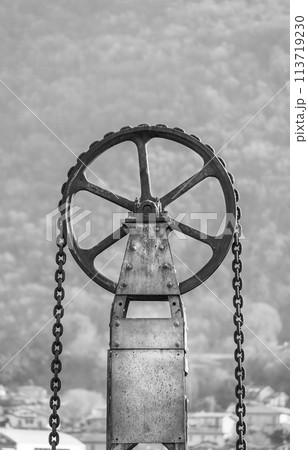 One round metal wheel of a lifting mechanism with a chain Black and white vertical photo 113719230