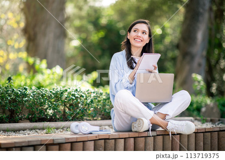 Thoughtful young woman with a laptop sitting cross-legged on a bench in a park Thoughtful young woman with a laptop sitting cross-legged on a bench in a park 113719375
