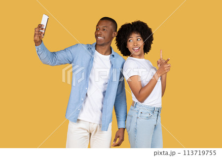 Happy African American man taking a selfie with his smartphone as the woman next to him points upwards Happy African American man taking a selfie with his smartphone as the woman next to him points upwards 113719755