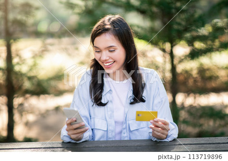Joyful young woman engaging with her smartphone while holding a credit card Joyful young woman engaging with her smartphone while holding a credit card 113719896