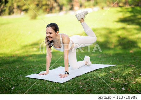 Smiling woman in white sportswear performs a donkey kick exercise on a yoga mat in a sunny park Smiling woman in white sportswear performs a donkey kick exercise on a yoga mat in a sunny park 113719988