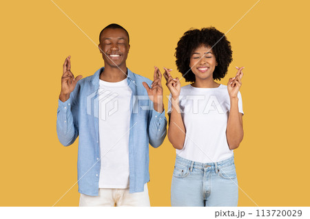 A cheerful young African American couple wearing casual clothing are crossing their fingers with eyes closed A cheerful young African American couple wearing casual clothing are crossing their fingers with eyes closed 113720029