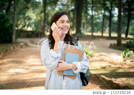Joyful young woman talking on a mobile phone while holding notebooks 113720216