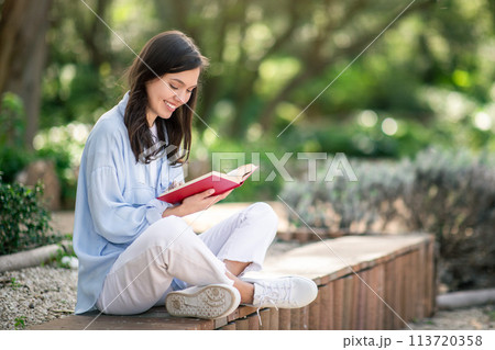 Serene young woman enjoying reading a red book, sitting cross-legged on a park bench Serene young woman enjoying reading a red book, sitting cross-legged on a park bench 113720358
