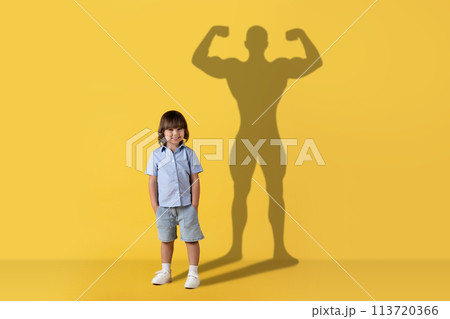 A cheerful young child stands smiling with a confident, muscular superhero shadow cast A cheerful young child stands smiling with a confident, muscular superhero shadow cast 113720366