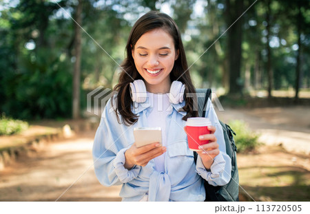 Contented young lady with headphones checking her smartphone and enjoying a warm beverage 113720505