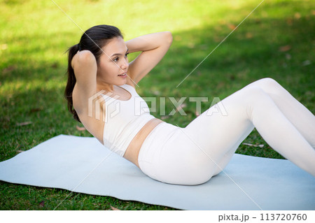 Focused woman in white activewear doing crunches on a yoga mat in a park Focused woman in white activewear doing crunches on a yoga mat in a park 113720760