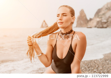 Woman travel portrait. Happy woman with long hair looking at camera and smiling. Close up portrait cute woman in black bikini posing on a red volcanic rock on the sea beach 113722999