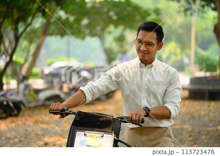 Smiling young businessman walking with bicycle through urban park 113723476