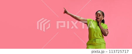 Banner. African-American woman looks surprised and pointing to side against pastel pink studio background with negative space. 113725717
