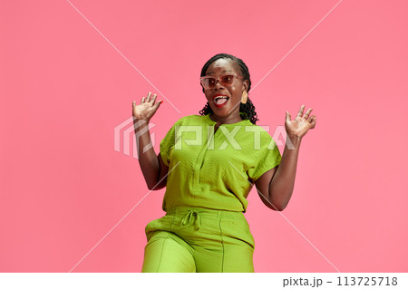 Existed young African-American woman in casual attire raising hands in delight against pastel pink studio background. 113725718