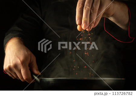 The cook adds aromatic spices to a hot, steamy frying pan. Low key concept of preparing a delicious dinner in a tavern kitchen. European cuisine The cook adds aromatic spices to a hot, steamy frying pan. Low key concept of preparing a delicious dinner in a tavern kitchen. European cuisine 113726782