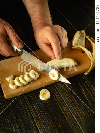 A cook uses a knife to slice a ripe banana to prepare a fruit dish on the kitchen table. Place for advertising on a dark background 113726785
