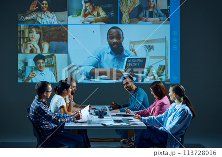 Workers sitting in office boardroom participate in virtual meeting, with remote colleagues via online video call 113728016