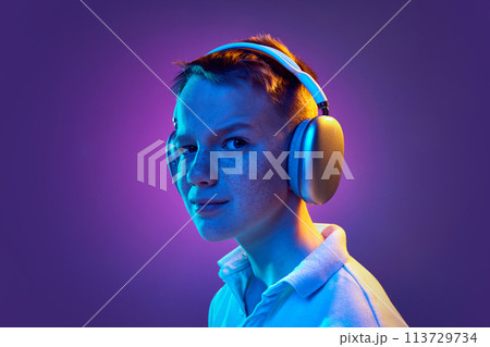 Portrait of young boy in headphones raising eyebrow looking at camera in neon light against purple gradient background. Portrait of young boy in headphones raising eyebrow looking at camera in neon light against purple gradient background. 113729734