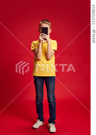 Full-length portrait of little boy using smartphone to take photo against red studio background. Full-length portrait of little boy using smartphone to take photo against red studio background. 113729813