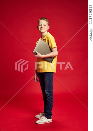 Full-length portrait of smiling school-boy in casual attire hold digital laptop against red studio background. 113729818
