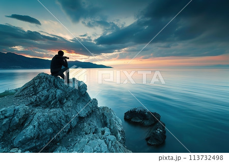 Professional Nature Photographer Photographer sits on a cliff looking at sunset sea. Travel adventure concept, active vacations outdoor. Rear view of traveler with his camera in hand on the rock top 113732498