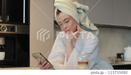 Portrait of happy woman wearing a bathrobe and a towel on her head using smartphone in the kitchen sending message on social media sharing. The morning is routine. Portrait of happy woman wearing a bathrobe and a towel on her head using smartphone in the kitchen sending message on social media sharing. The morning is routine. 113735092