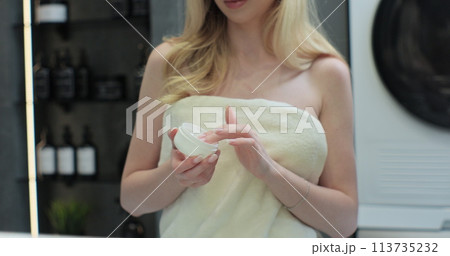 Woman wrapped in bath towel putting cream on face, smiling, looking at mirror. Concept of skincare routine. Skincare and bodycare. 113735232
