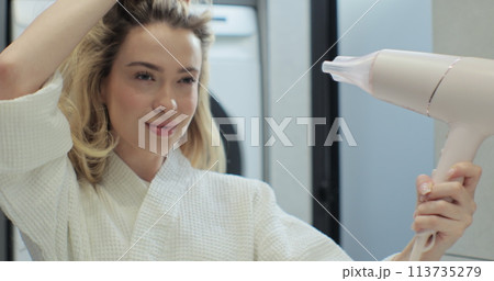 Close up of attractive young woman looking at her reflection in the mirror and drying hair with blow dryer in bathroom. Concept of well-groomed and beautiful woman. 113735279