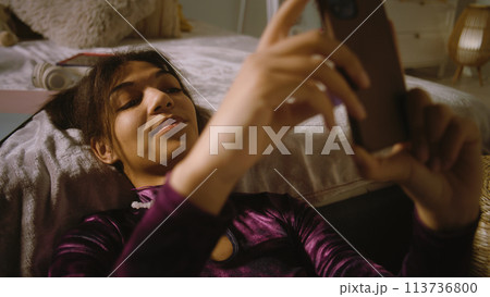 Girl sits near bed, smiles and surfs internet using phone Girl sits near bed, smiles and surfs internet using phone 113736800