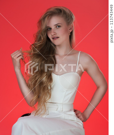 Portrait of young blonde woman dressed in white sculpted corset top. Headshot of young female fashion model from Generation Z with long, wavy blonde hair looking at camera against red background Portrait of young blonde woman dressed in white sculpted corset top. Headshot of young female fashion model from Generation Z with long, wavy blonde hair looking at camera against red background 113743449