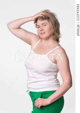 Woman with plump figure poses with one hand raised to head and other hand in pocket, standing on white background. Authentic mature model looking at camera, wearing camisole and green jogging pants 113743482