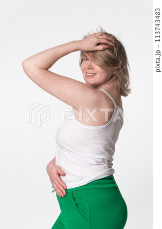 Portrait of happy plump woman is smiling and looking over her shoulder at camera, posing with one hand raised to head and hand on stomach. She wearing camisole and green sweatpants on white background 113743483