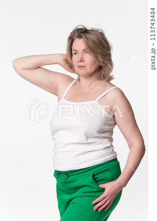 Portrait of chubby mature adult blonde woman Generation X model wearing cotton camisole and green jogging pants. Comely woman with one hand raised behind head looking away, posing on white background 113743484
