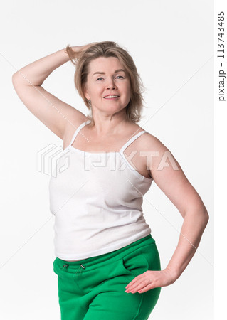 Portrait of happy plump mature adult blonde woman with medium length hair. Smiling model with one hand raised behind head wearing white cotton camisole and green sweatpants, posing on white background 113743485