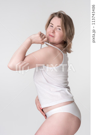 Woman wearing underwear holding strap of camisole and placing hand on stomach. Representative of concept of healthy lifestyle for women of Generation X Caucasian demographic, regardless of body type 113743486