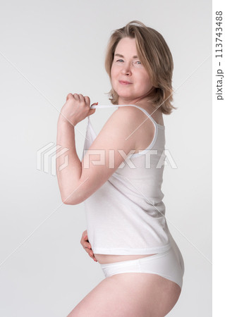 Middle aged overweight woman wearing white underwear, holding strap of camisole and placing hand on her stomach. Caucasian Generation X woman represents the concept of a healthy lifestyle for ladies 113743488