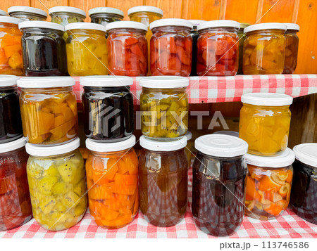Assortment of homemade preserves in glass jars with checkered cloth, various pickled fruits and vegetables, colorful display, canned goods collection, close up with selective focus. 113746586