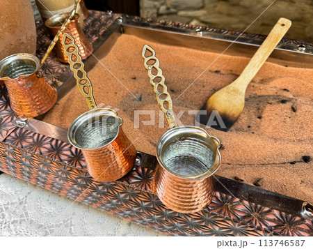 Traditional Turkish coffee in copper turks with long handles on tray with sand. Real coffee preparation, close up with selective focus. 113746587