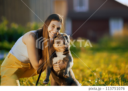 Trying to force dog to look into camera lens, young white woman takes photo with an American Bully in backyard of country house on summer evening. 113751076