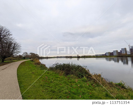 London, UK - 12.16.2021: An empty pathway next to Low Maynard Reservoir in the Walthamstow Wetlands along trees and marshes under a cloudy sky with buildings at the edge, viewed from Mayward Entrance London, UK - 12.16.2021: An empty pathway next to Low Maynard Reservoir in the Walthamstow Wetlands along trees and marshes under a cloudy sky with buildings at the edge, viewed from Mayward Entrance 113751354