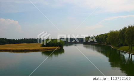 Zhujiajiao Ancient town, Shanghai, China - July 25, 2019: peaceful lake surrounded by willow trees and grassland with a wooden bridge in the background under a bright blue sky 113752169