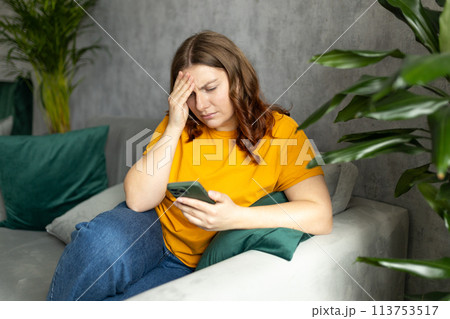 Portrait of beautiful young woman with depressed facial expression sitting on the couch holding her phone. Sad 30s women desperately looking at smartphone screen 113753517
