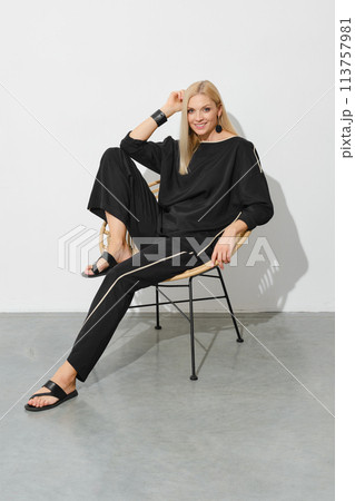 Smiling woman in loose casual clothes and leather sandals sits in profile on a chair Smiling woman in loose casual clothes and leather sandals sits in profile on a chair 113757981