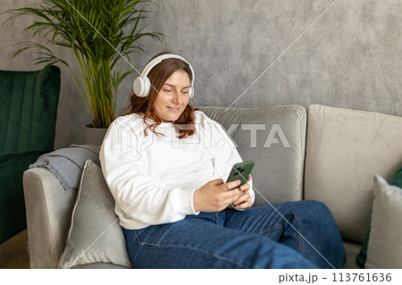 Smiling girl relaxing at home, she is listening to music, using a smartphone and wearing white headphones. Young 30s woman hearing enjoying good audio sound lounge on couch at home, full body 113761636