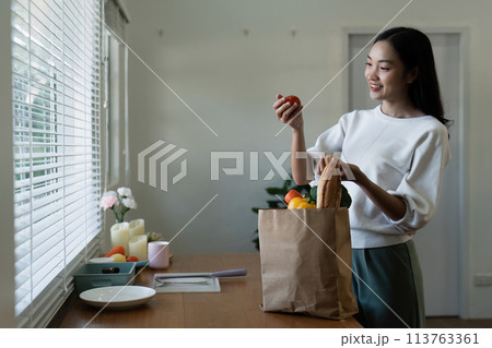 Organic Food Delivery. Happy young woman unpacking bag with Fresh Vegetables in kitchen Organic Food Delivery. Happy young woman unpacking bag with Fresh Vegetables in kitchen 113763361