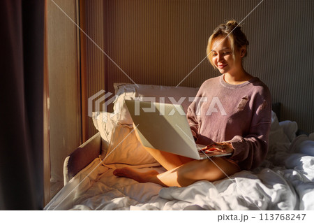 Happy casual beautiful woman working on a laptop sitting on the bed in the house. Happy casual beautiful woman working on a laptop sitting on the bed in the house. 113768247