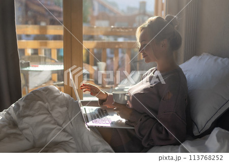 Happy casual beautiful woman working on a laptop sitting on the bed in the house. Happy casual beautiful woman working on a laptop sitting on the bed in the house. 113768252