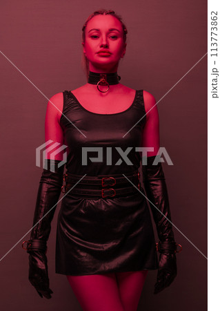 Beautiful young woman in a leather dress and bondage set posing on red light backgound Beautiful young woman in a leather dress and bondage set posing on red light backgound 113773862