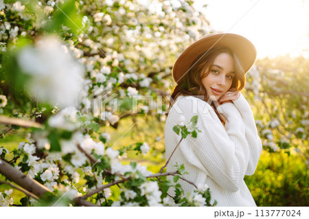 Portrait of Woman In hat posing near flowering tree. Smiling young woman enjoying smell of flowers. 113777024