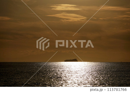 夕暮れの太陽光が海に反射して水平線に浮かび上がる貨物船のシルエット 113781766