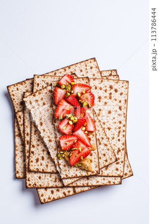 Matza toast with peanut butter, strawberries and pistachios on a white plate. Traditional bread for the Jewish holiday of Passover. 113786444