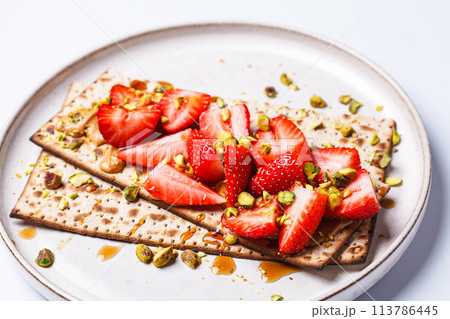 Matza toast with peanut butter, strawberries and pistachios on a white plate. Traditional bread for the Jewish holiday of Passover. 113786445
