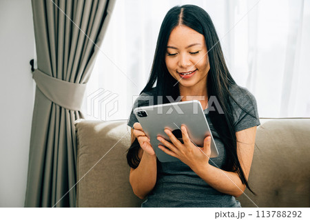Amidst a comfortable home atmosphere a blissful young Asian woman enjoys leisure time engaged in online shopping through a digital tablet on the sofa exuding relaxation and happiness. 113788292